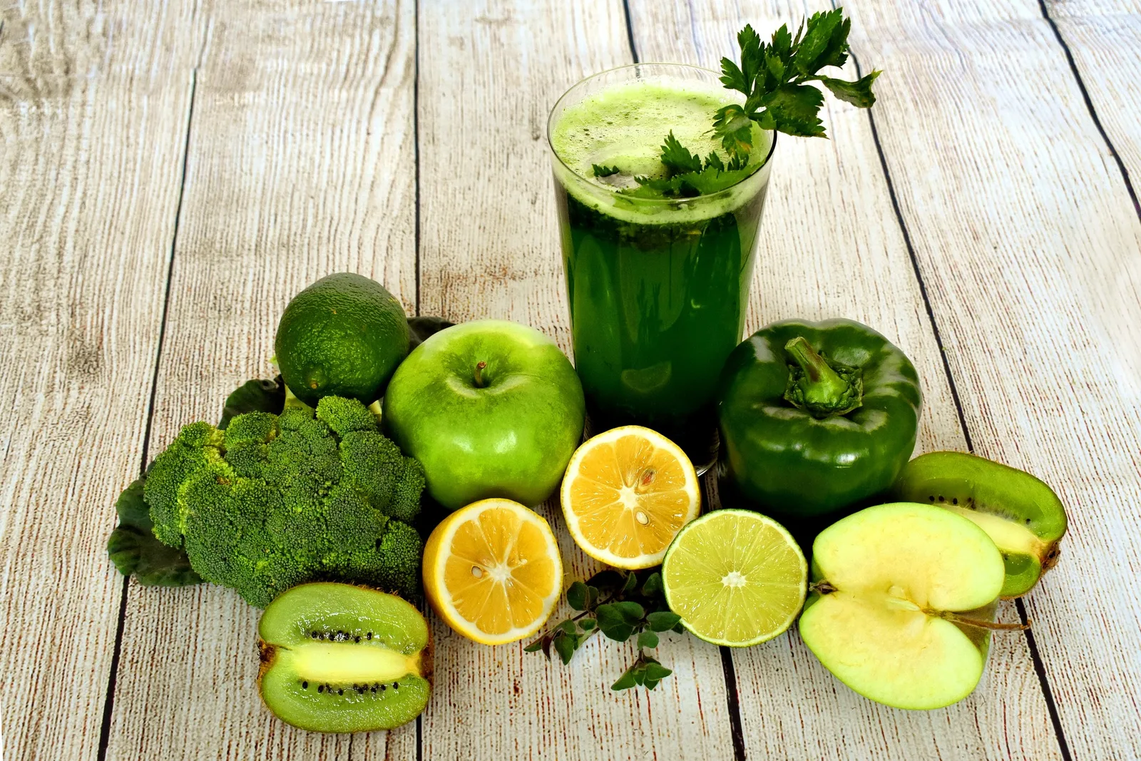 Glas mit grünem Smoothie umgeben von grünem Gemüse, Obst und Kräutern auf Holzuntergrund