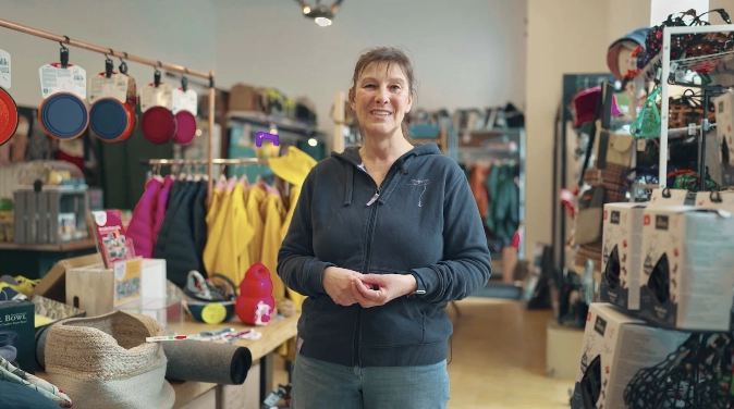 Frau in dunkler Jacke steht in Handwerkswerkstatt mit bunten Kochutensilien und Materialien im Hintergrund