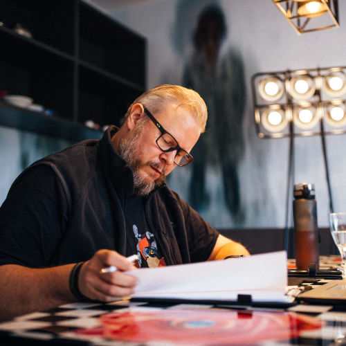 Mann mit Brille und schwarzem Shirt schreibt konzentriert an Schreibtisch mit Studiolampen im Hintergrund