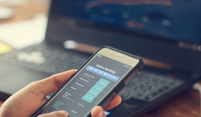Hand hält Smartphone mit Online-Banking-Interface, Laptop im Hintergrund