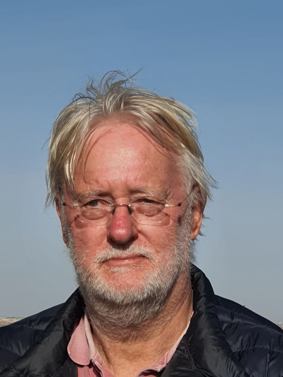 Outdoor-Porträt eines älteren Mannes mit weißen Haaren, Brille, schwarzer Jacke und rosa Hemd vor blauem Himmel