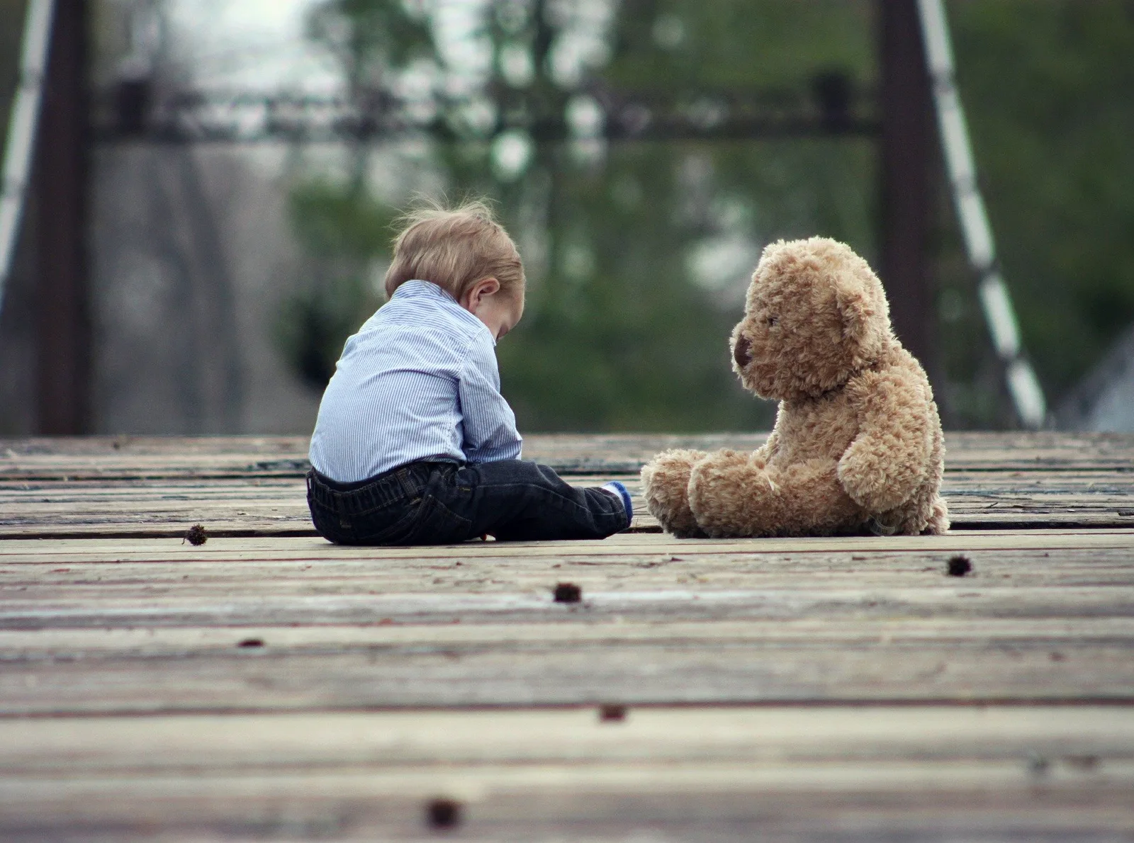 Kleines Kind sitzt auf Holzsteg neben Teddybär, grüne Brücke verschwommen im Hintergrund