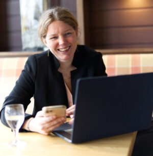 Patricia Junk lachend am Laptop mit Smartphone, schwarzer Blazer, Indoor-Setting