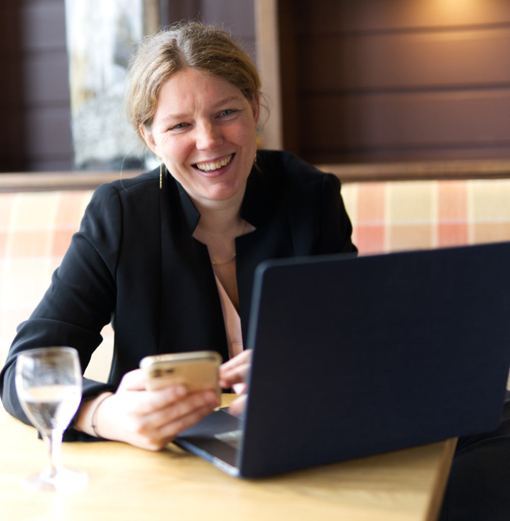 Patricia Junk lachend am Laptop mit Smartphone, schwarzer Blazer, Indoor-Setting