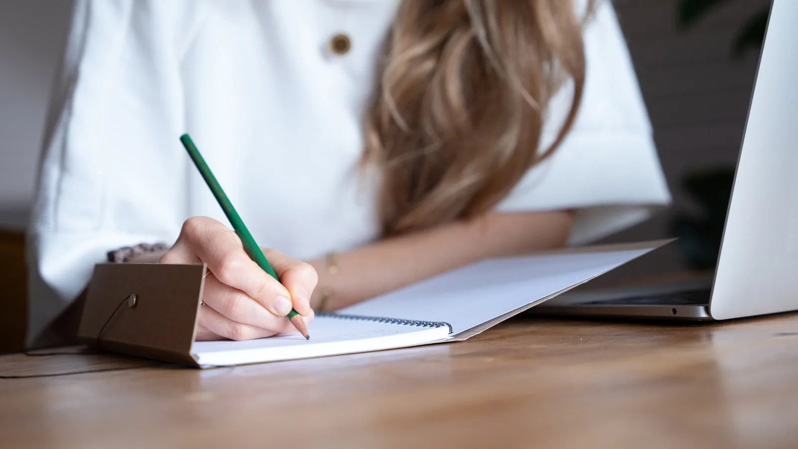 Frau in weißem Hemd schreibt mit grünem Stift in Notizbuch am Schreibtisch