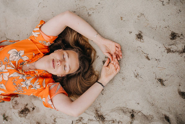Junge Frau in orangenem Blütenkleid liegt auf Sand am Strand