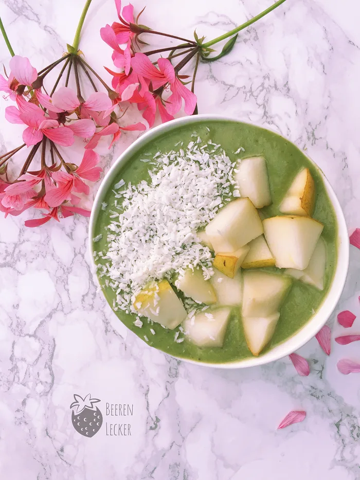 Grüne Gerstengras-Smoothie-Bowl mit Kokosflocken und Apfelstücken, dekoriert mit rosa Blüten