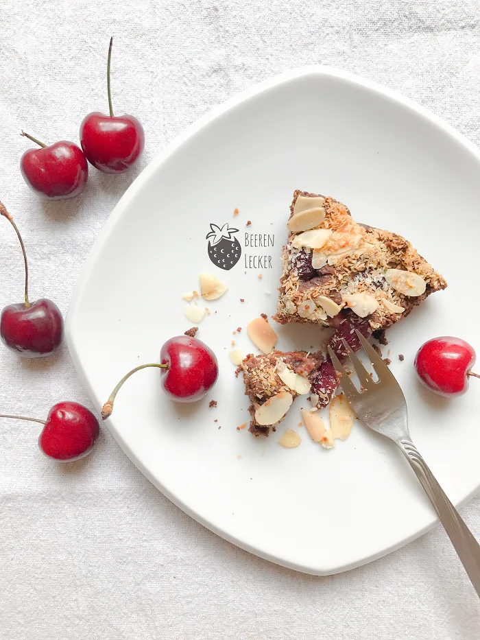 Stück veganer Kirsch-Schoko-Kuchen auf weißem Teller mit Mandeln, rote Kirschen daneben