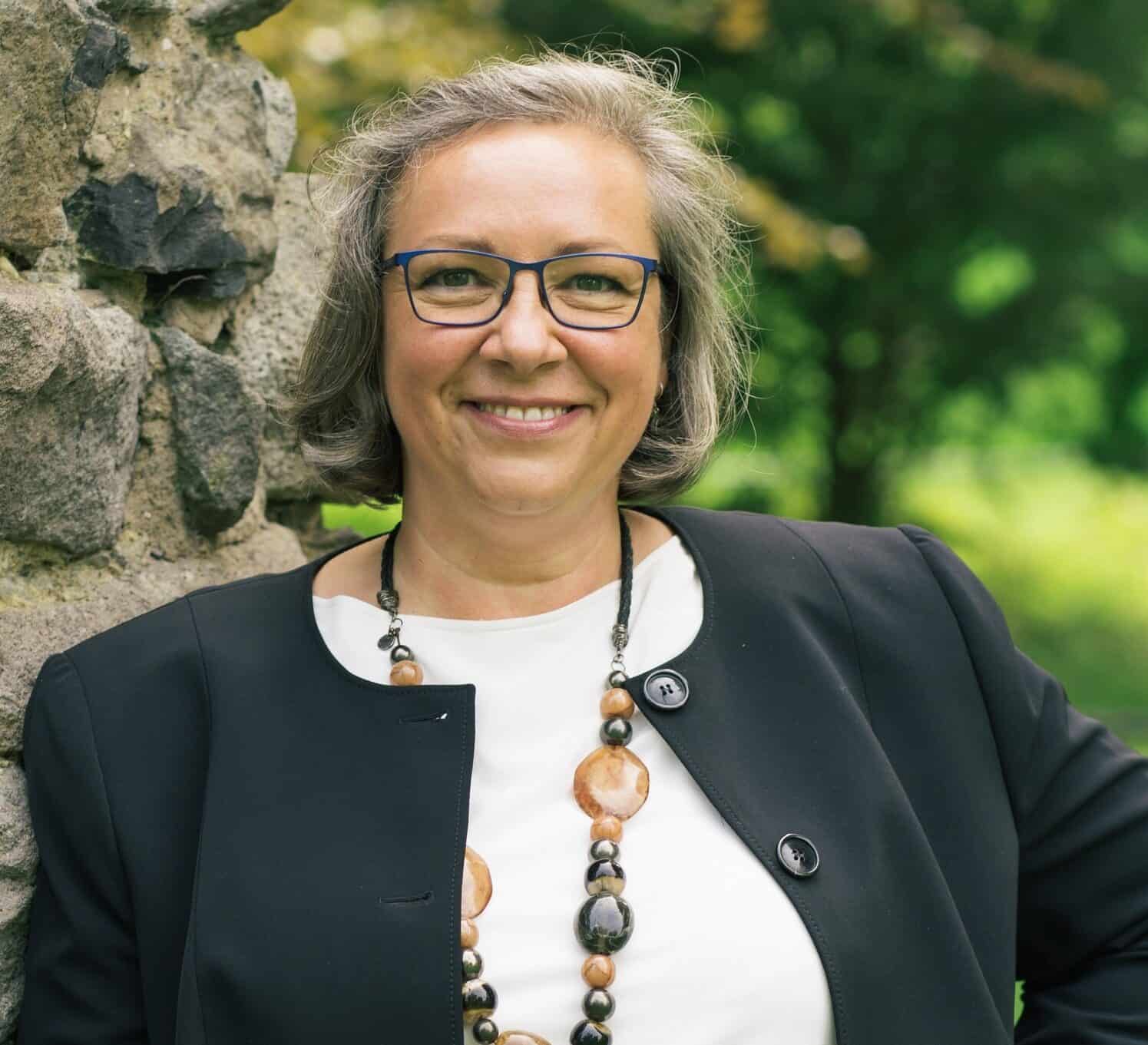 Porträt von Katrin Schikat mit Brille, schwarzem Blazer und dekorativem Schmuck