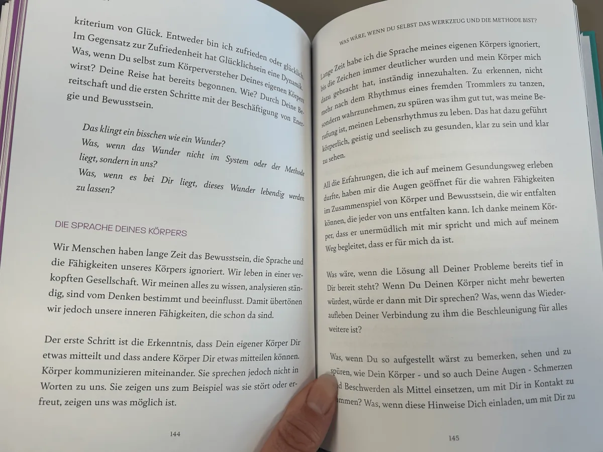 Aufgeschlagene Buchseiten 144-145 mit Text zu Glück und Körpersprache, Hand hält das Buch