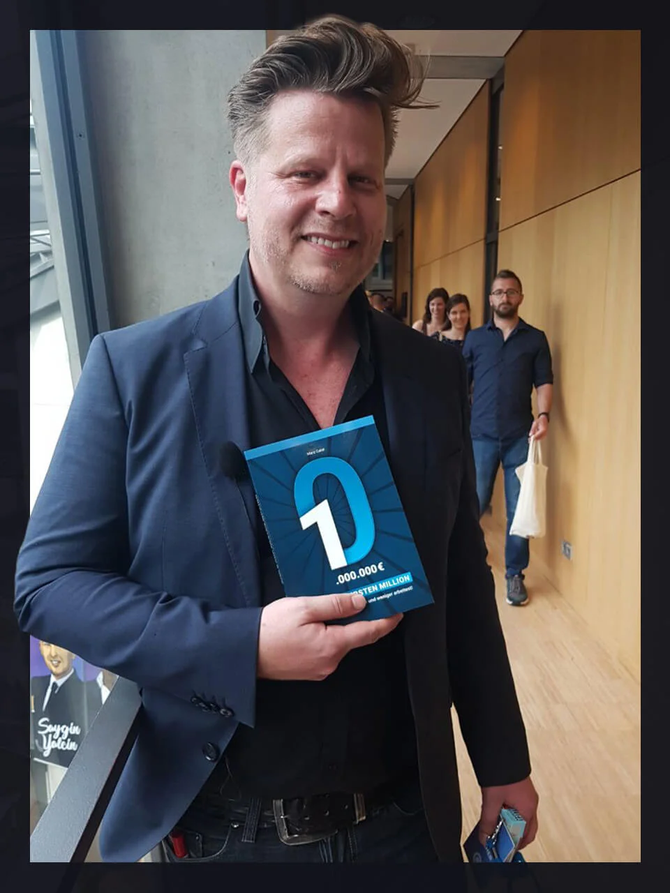 Lächelnder Mann im dunkelblauen Blazer hält blaues D-Hörbuch-Cover in Flur