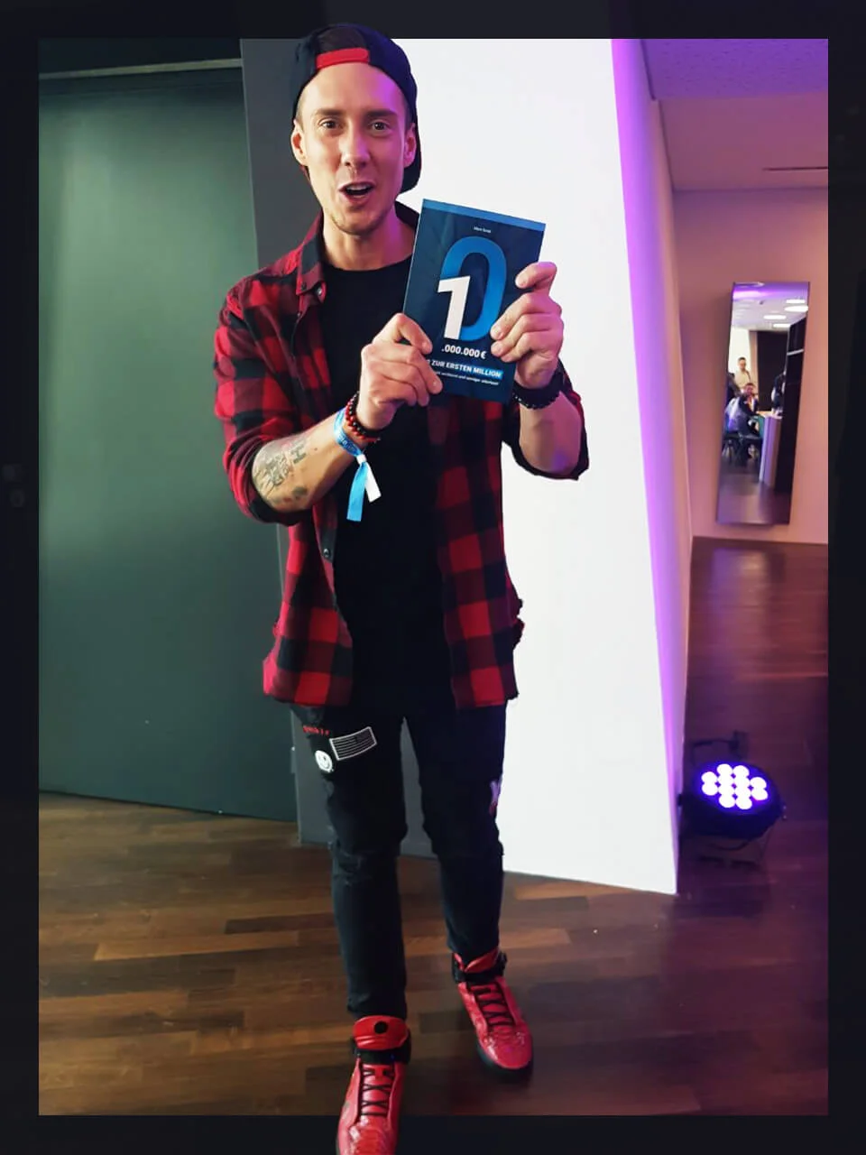 Mann mit rokarierter Kappe hält blaues Hörbuch-Cover mit großem D-Logo und Millionen-Text