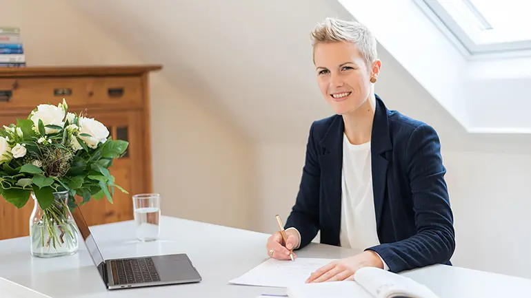 Frau in dunklem Blazer sitzt am weißen Schreibtisch und macht Notizen