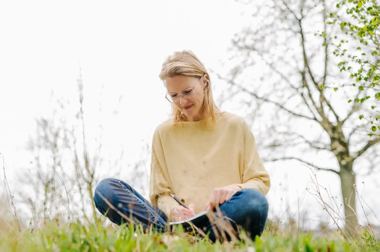 Frau im gelben Pullover sitzt nachdenklich in Natur auf Gras mit Tablet
