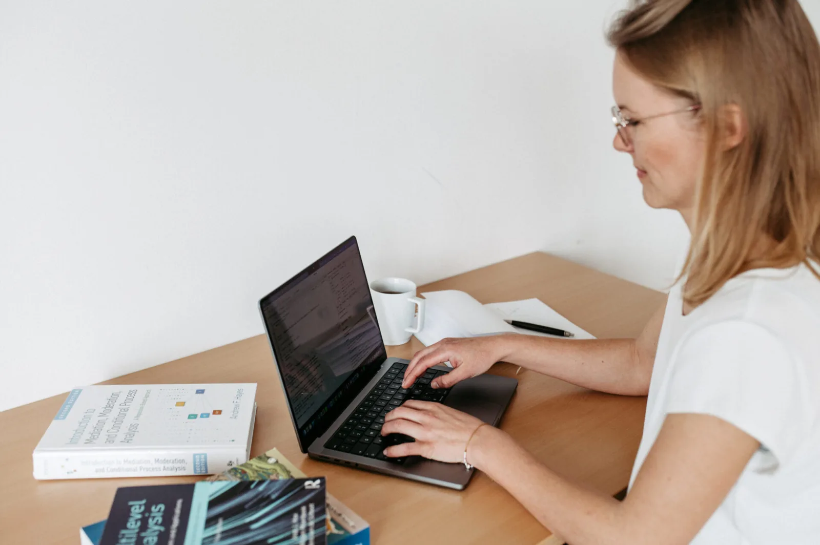 Frau mit Brille arbeitet am Laptop an einem Schreibtisch mit Kaffee und Lernmaterialien