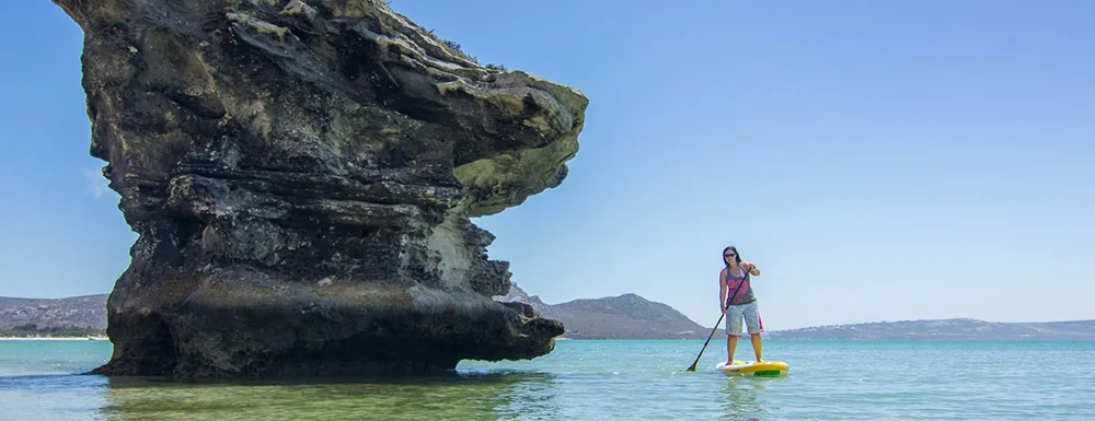 Stand-up-Paddlerin auf gelbem SUP-Board vor Felsen in Kraalbai Südafrika