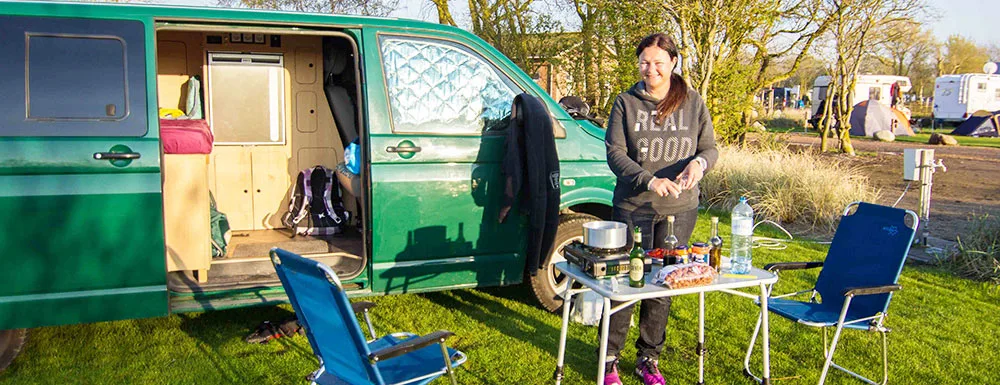 Frau kocht am Campingplatz neben grünem VW Bus auf Fehmarn