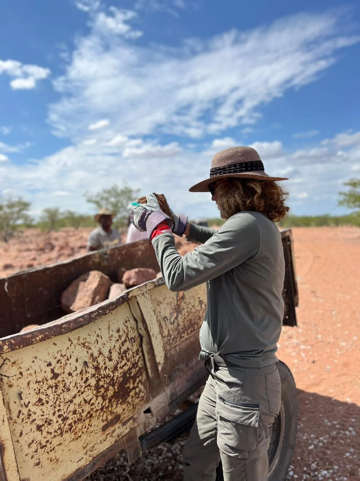 Cornelia Schumacher sammelt rote Steine in Australien-Outback, trägt Strohhut und Arbeitshandschuhe, Traktor im Hintergrund