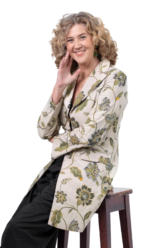Cornelia Schumacher sitzt auf Stuhl, trägt Blazer mit Blüten-Muster, hand am Gesicht, lächelt zur Kamera