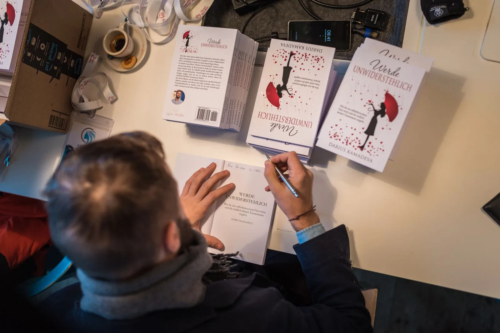 Person signiert Buch "Werde unwiderstehlich" von Darius Kamadeva auf weißem Tisch mit mehreren Exemplaren