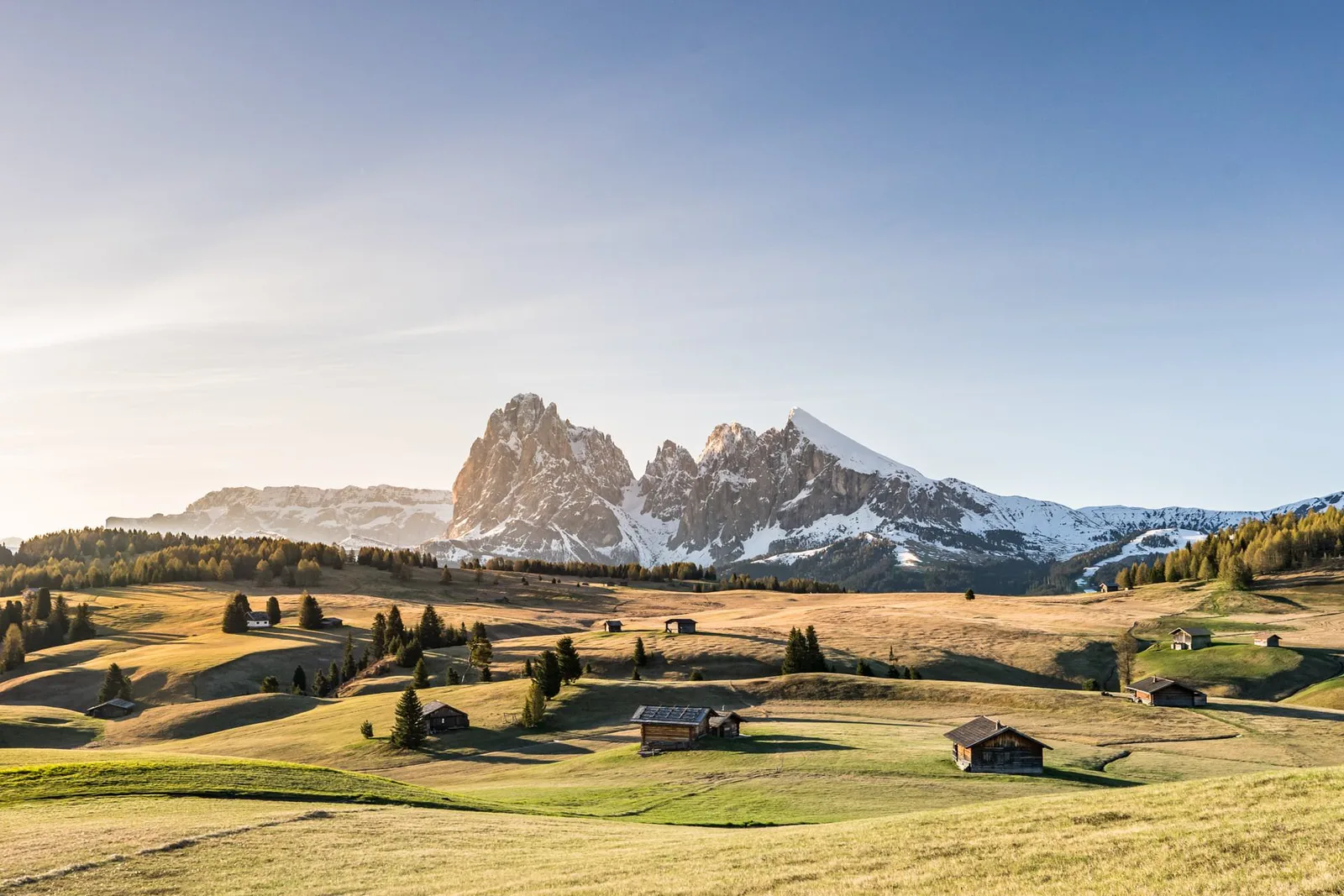 Berglandschaft mit schneebedeckten Gipfeln, grünen Wiesen und traditionellen Almhütten in den Dolomiten