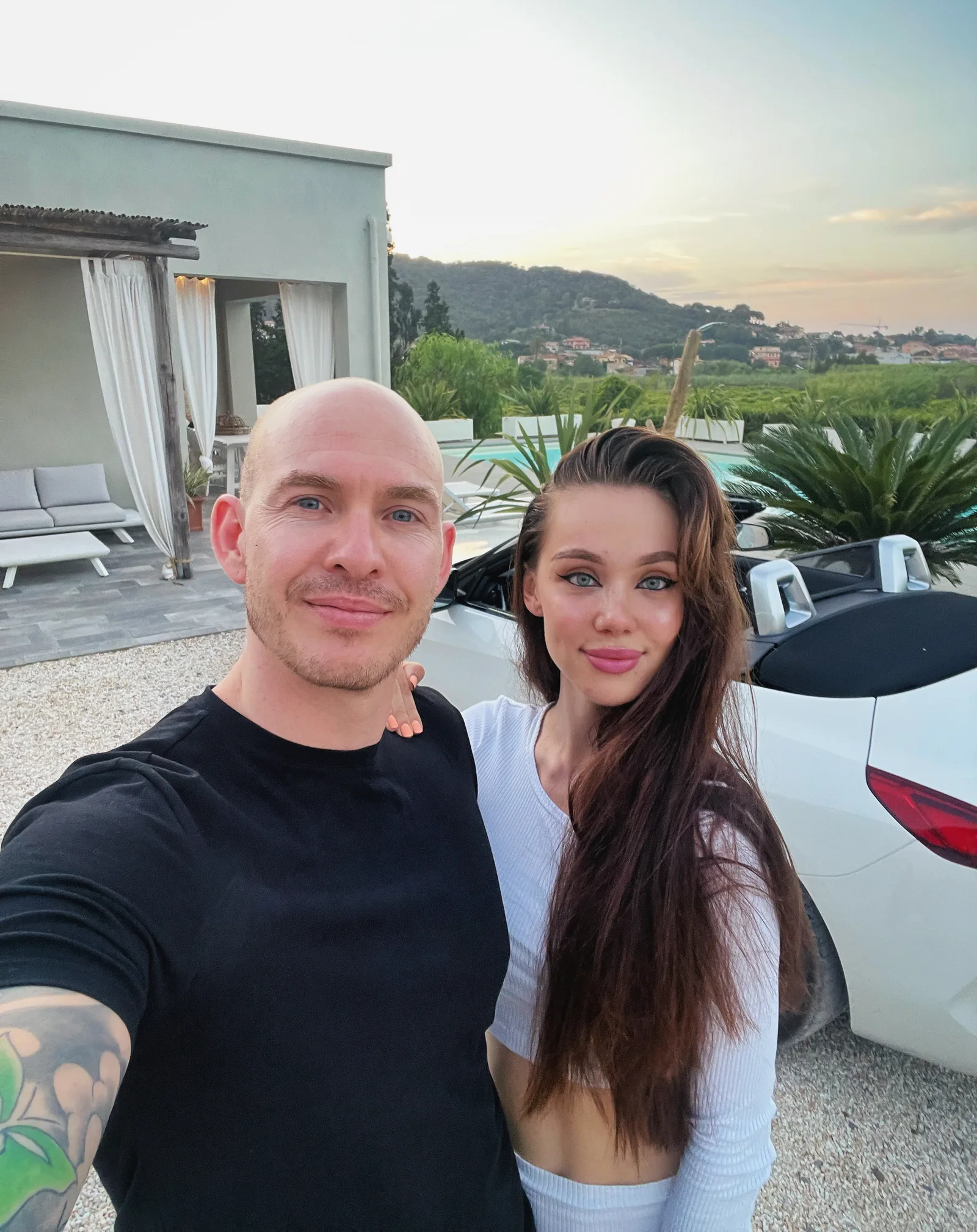 Mann und Frau vor Luxusvilla mit Pool, bergige Landschaft im Hintergrund, weißer Wagen