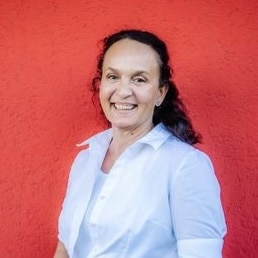 Porträtfoto von Karin Forstner, Frau mit dunklen Haaren in hellblauem Oberteil vor rotem Hintergrund