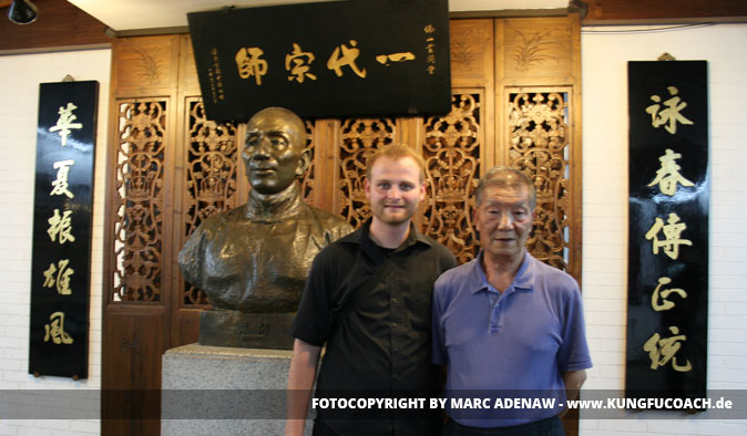 Mann und erfahrener Trainer vor chinesischer Bronzestatue und traditionellen Wandtafeln mit Schriftzeichen