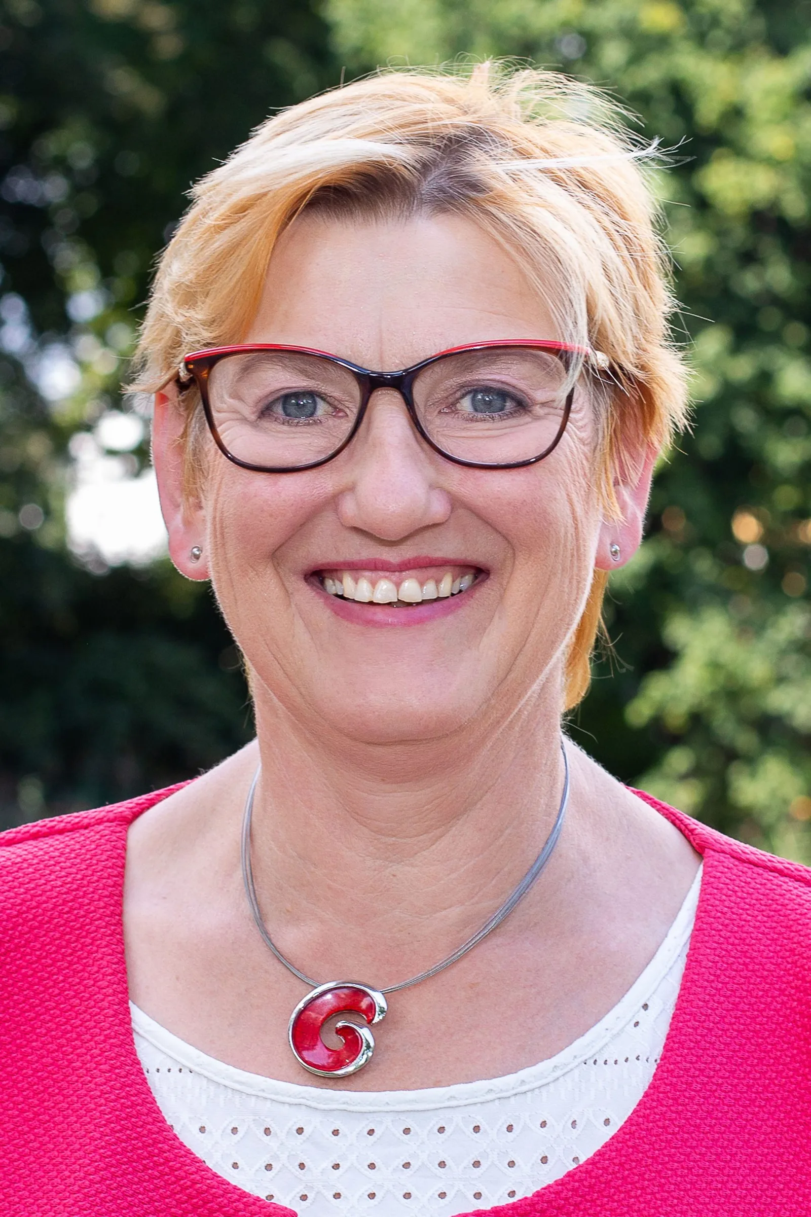 Wirtschaftskongress Vogtland – Video-Aufzeichnung von Nadine Täubert 10 Frau mit rötlichen Haaren, Brille, rosa Pullover mit rotem Anhänger und lächelndem Ausdruck vor grünem Hintergrund