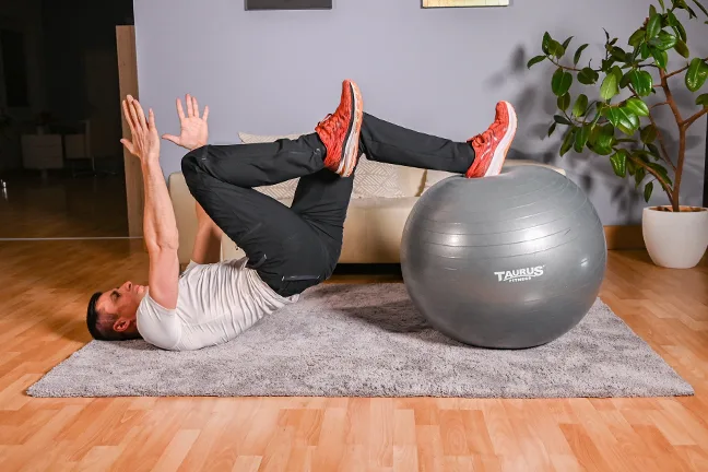 Mann beim Stabilitätstraining mit grauem Fitnessball auf Matte im Wohnzimmer