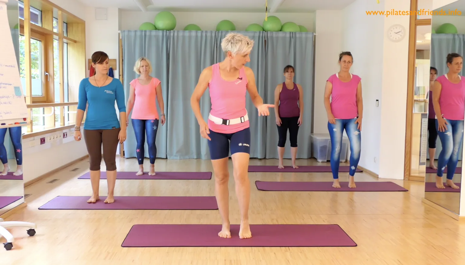 Pilates-Trainerin in rosa demonstriert Übung vor Gruppe auf violetten Matten