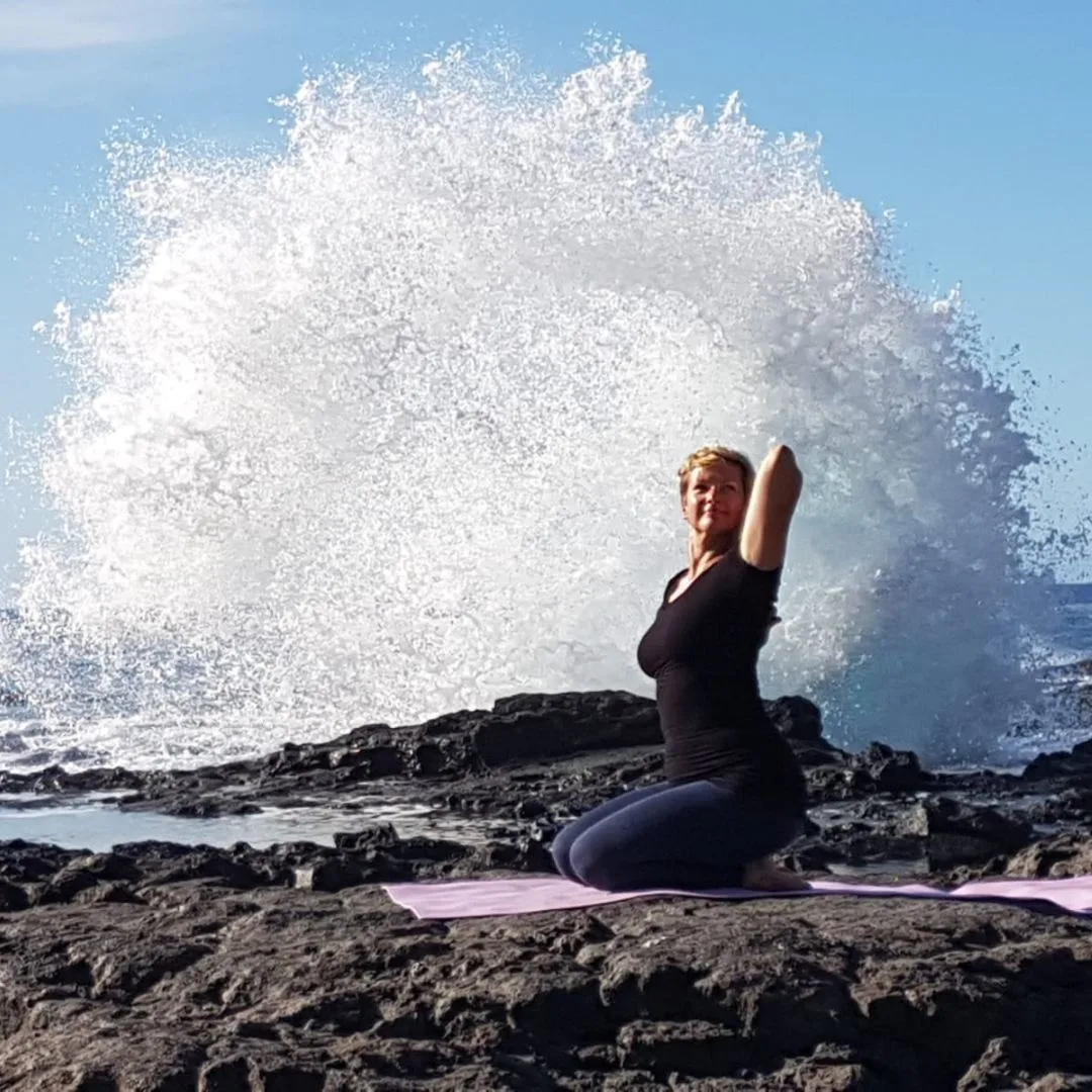 Yoga Live Online Kurs von Anke Schneider 1 Frau in schwarzer Sportbekleidung praktiziert Yoga-Pose auf lila Matte am Meer mit großer Wasserwelle im Hintergrund