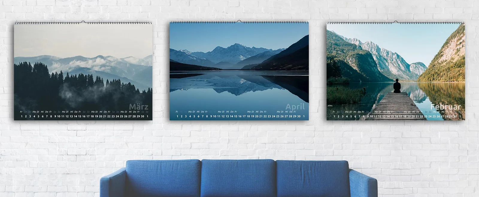 Drei YouDesign Calendar Wandkalender an weißer Backsteinwand mit Berglandschaften und Monatsnamen