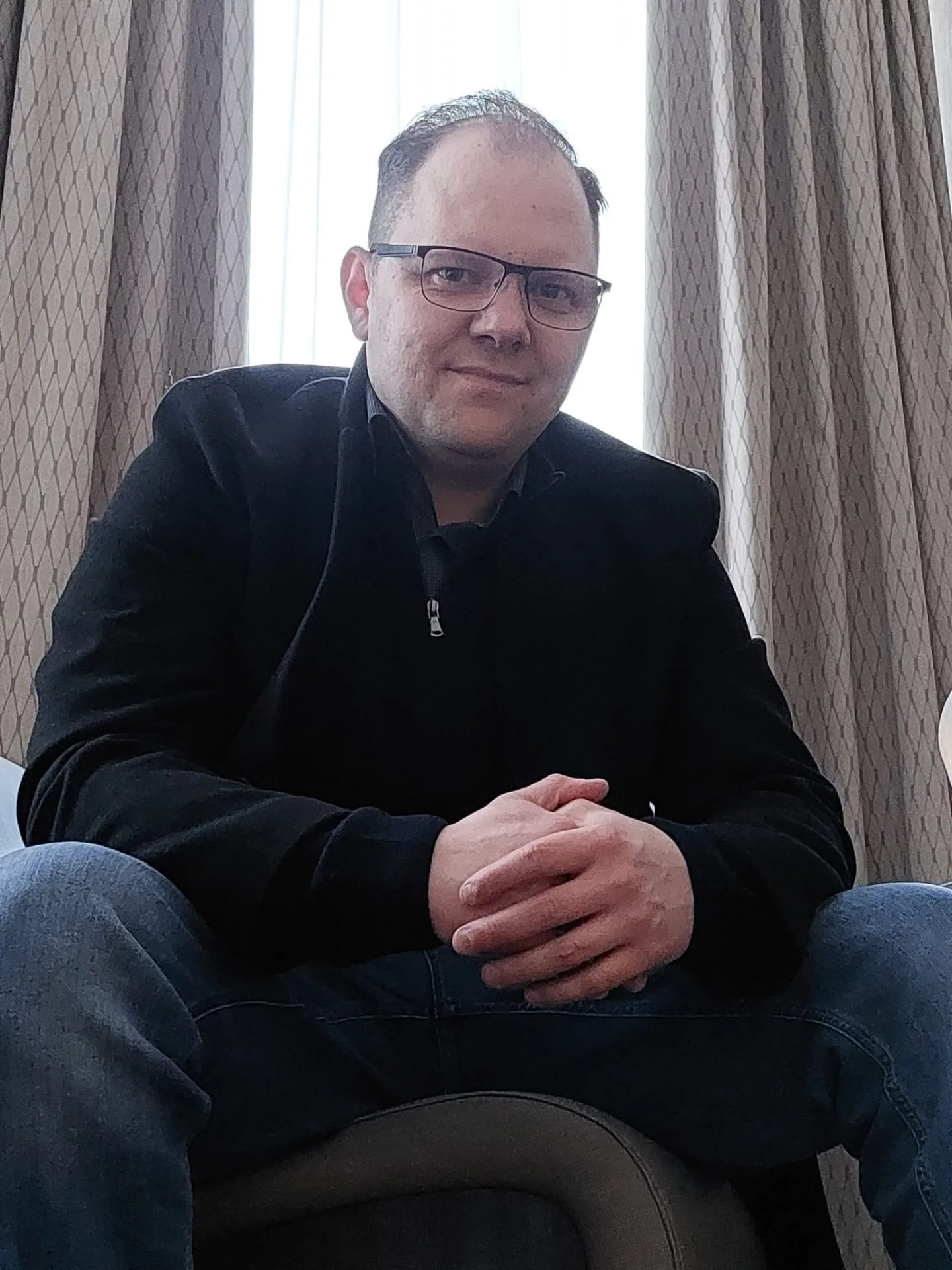 Portrait eines Mannes mit Brille in schwarzem Oberteil, sitzend vor Vorhang