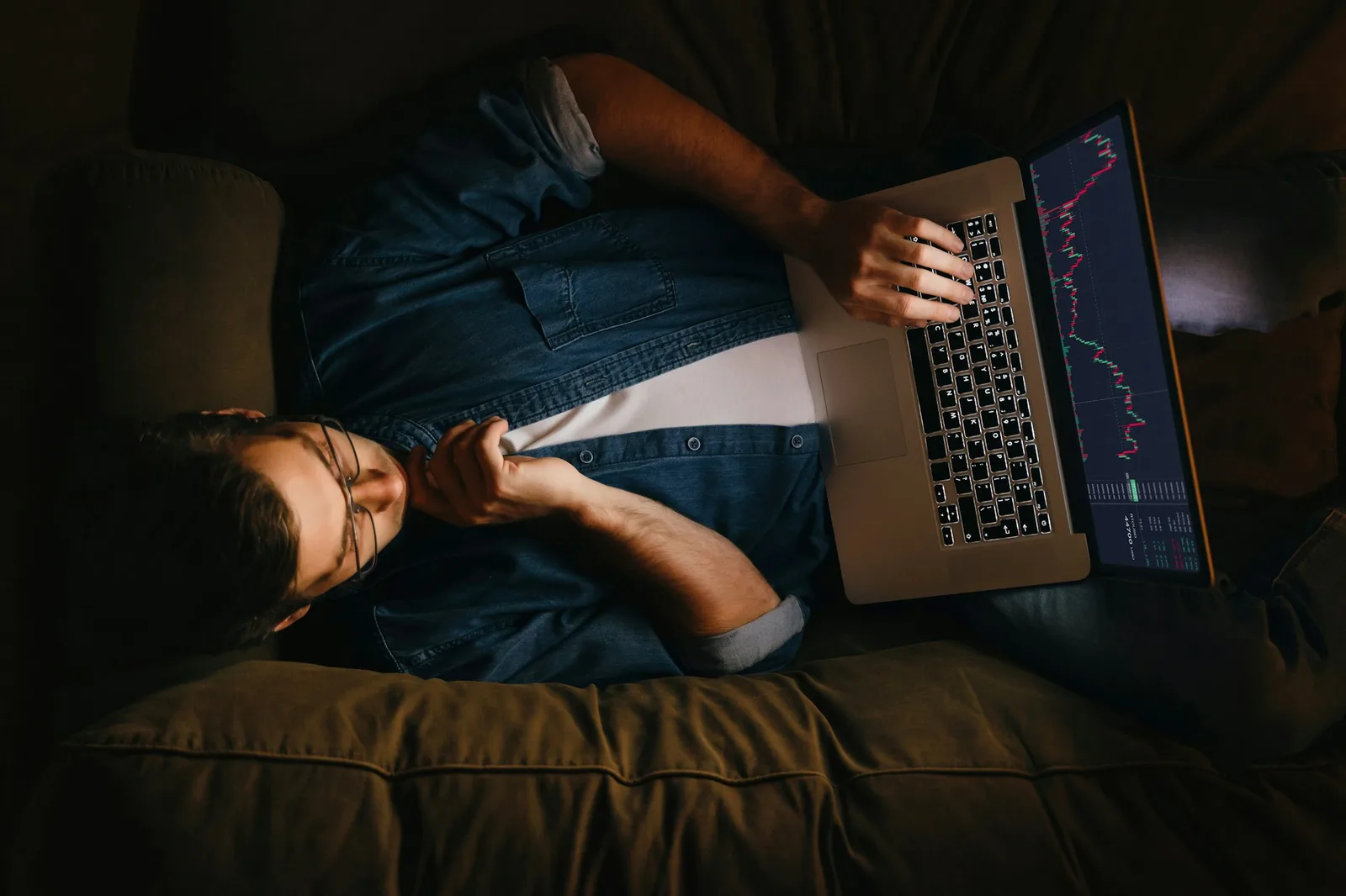 Junger Trader sitzt auf Bett und schaut auf Laptop-Bildschirm mit Börsendiagrammen