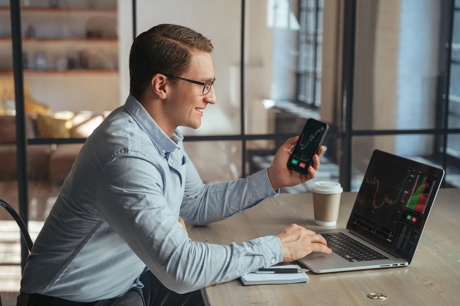 Mann mit Brille schaut lächelnd auf Smartphone, Laptop mit Candlestick-Charts im Hintergrund