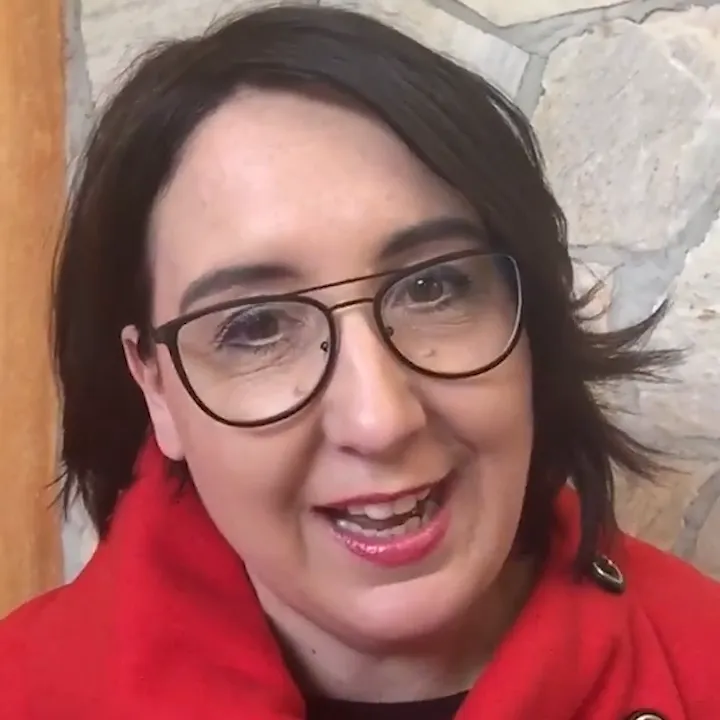 Frau mit Brille, rotem Shirt und dunklem Bob-Haarschnitt lächelnd vor Steinwand