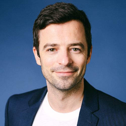 Porträtfoto von Manuel Burzler, Mann mit dunklem Haar, dunklem Blazer vor blauem Hintergrund