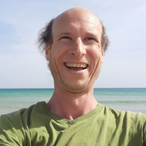 Alexander Seelendank, Mann mit kahlem Kopf lächelnd vor hellblauem Hintergrund am Strand