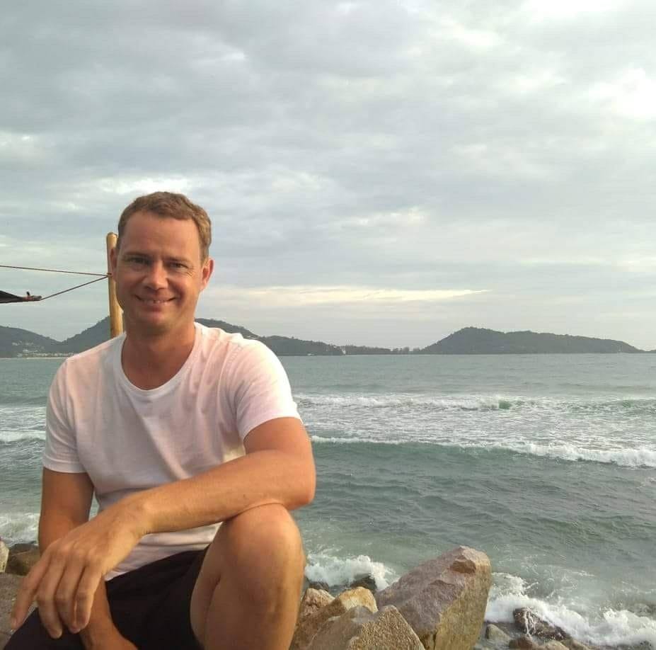 Mann im weißen T-Shirt lächelt in die Kamera, sitzt auf Felsen am Meer mit Insellandschaft