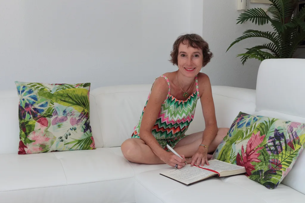 Isabel Ihm sitzt auf Sofa mit Notizbuch, buntes Muster-Tankshirt, grüne Pflanze, bunte Deko-Kissen