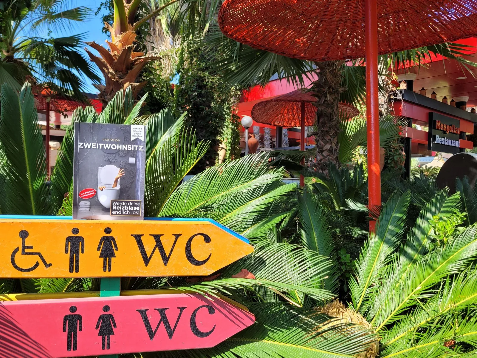 Zweitwohnsitz Buch in tropischem Restaurant-Ambiente bei WC-Hinweis