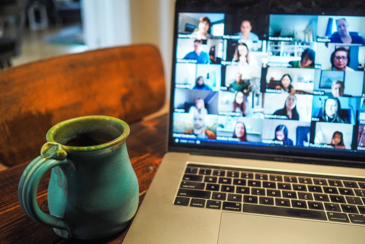 Laptop-Bildschirm mit Video-Konferenz zeigt mehrere Teilnehmer, daneben grüne Kaffeetasse auf Holztisch