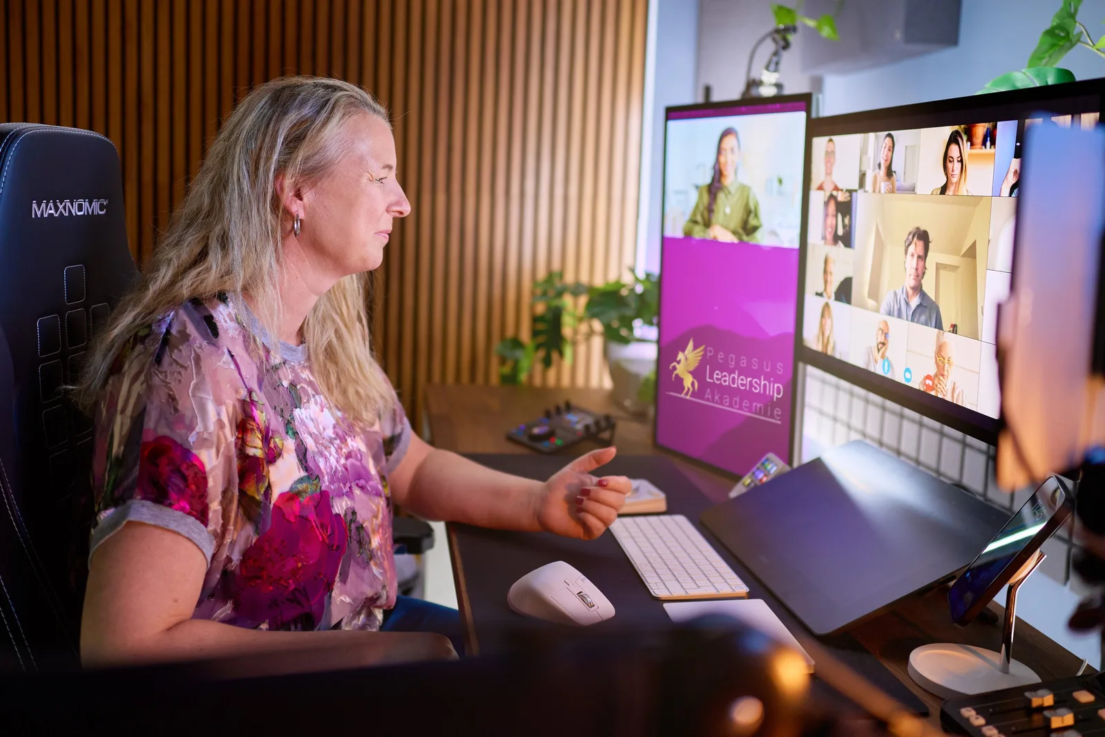 Frau mit blonden Haaren sitzt am Schreibtisch vor Monitor mit Pegasus Leadership Akademie Logo und Videokonferenz-Teilnehmern