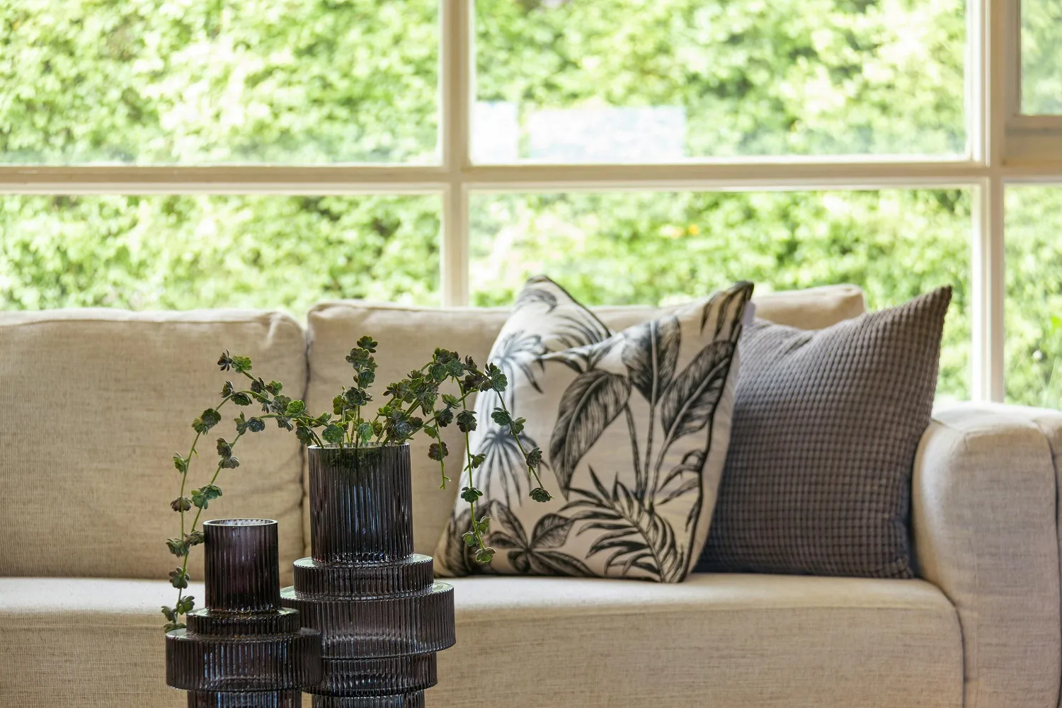 Helles Wohnzimmer mit Sofa, Kissen und Grünpflanzen vor großem Fenster