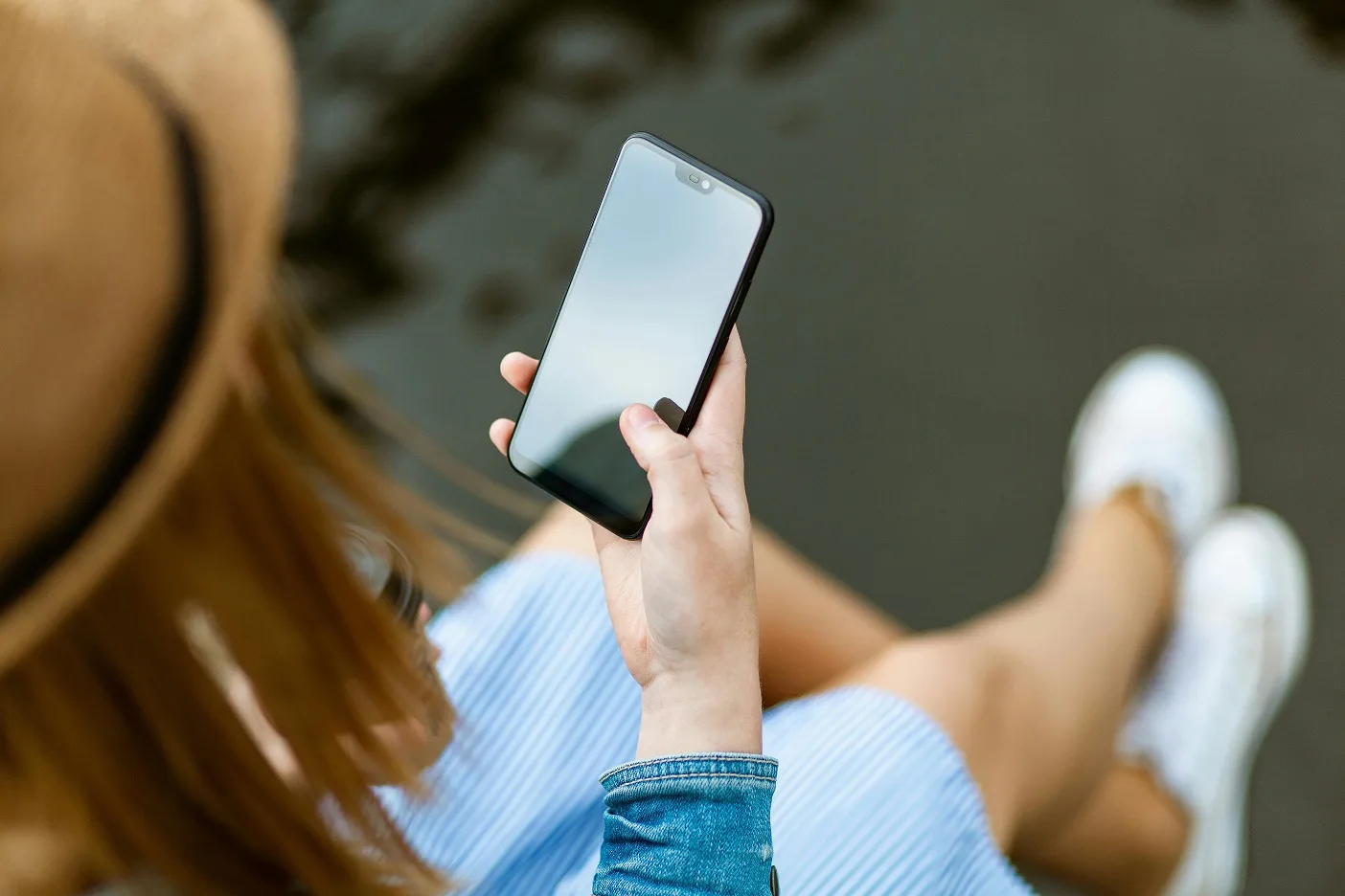 Frau von hinten hält Smartphone in Händen, trägt blaues Shirt und weiße Sneaker