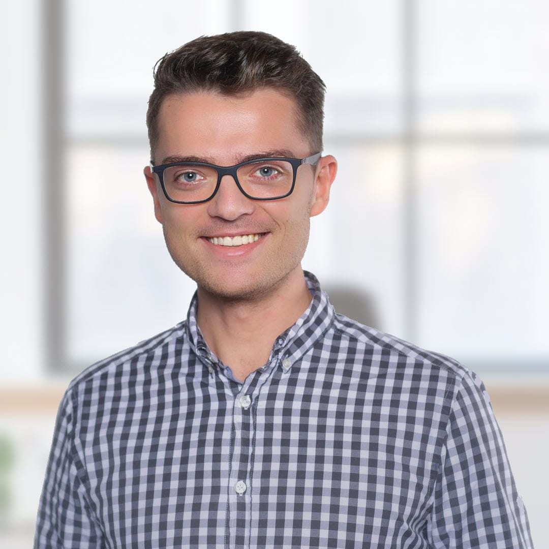 Porträtfoto eines lächelnden Mannes mit Brille in kariertem Hemd vor unscharfem Büro-Hintergrund