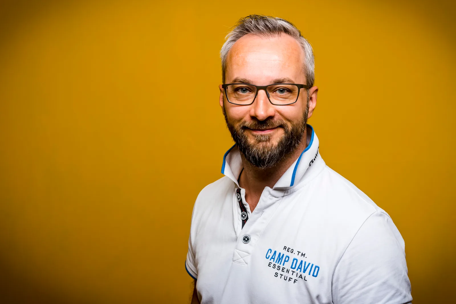 Christian Enengl, Mann mit Brille und Bart in weißem Polo-Shirt vor gelbem Hintergrund