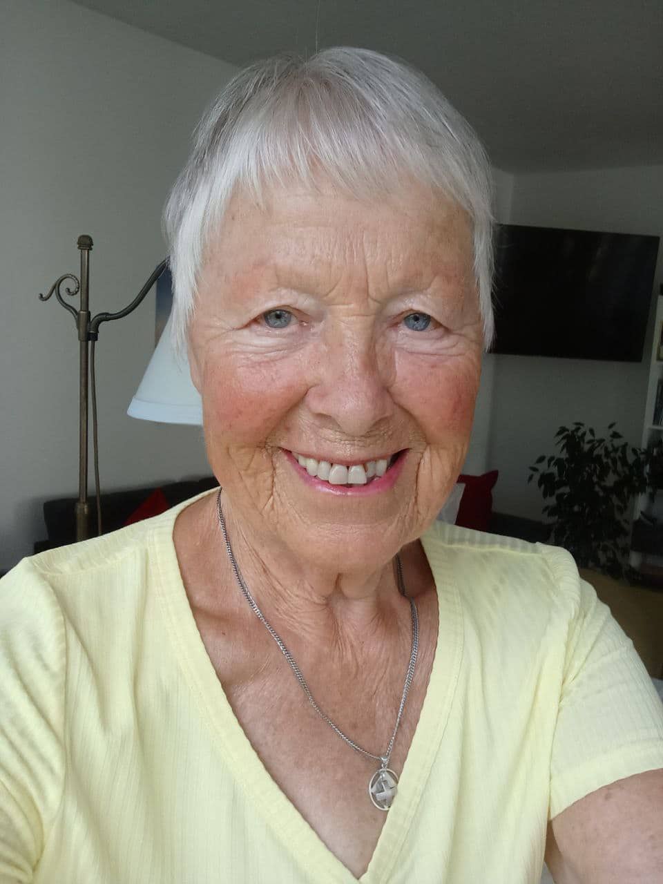 Ältere Frau mit weißen Haaren, gelbem Oberteil und Halskette lächelnd im Innenraum