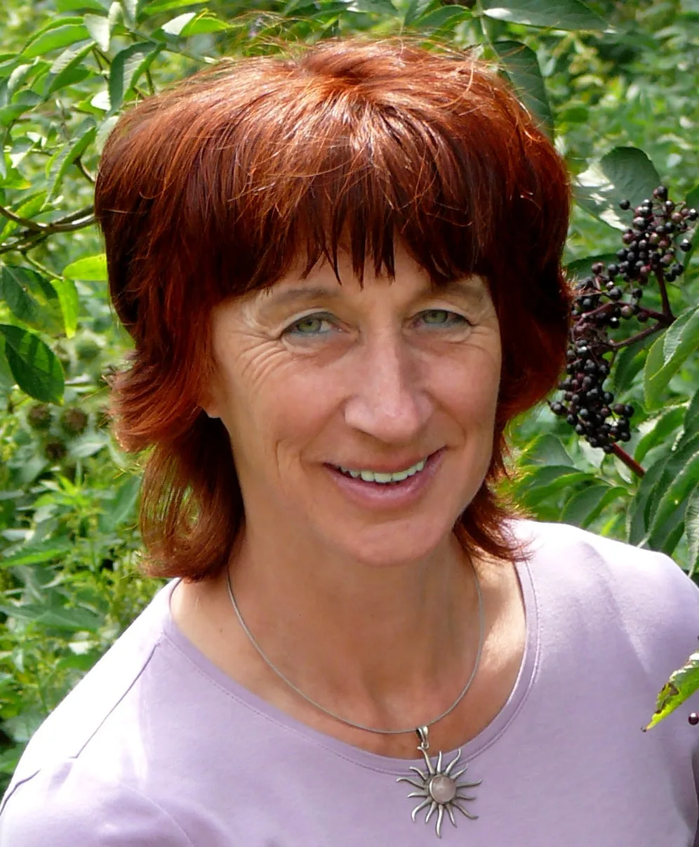 Ellen Huber, Frau mit roten Haaren und Halskette vor grünem Blätter-Hintergrund lächelnd